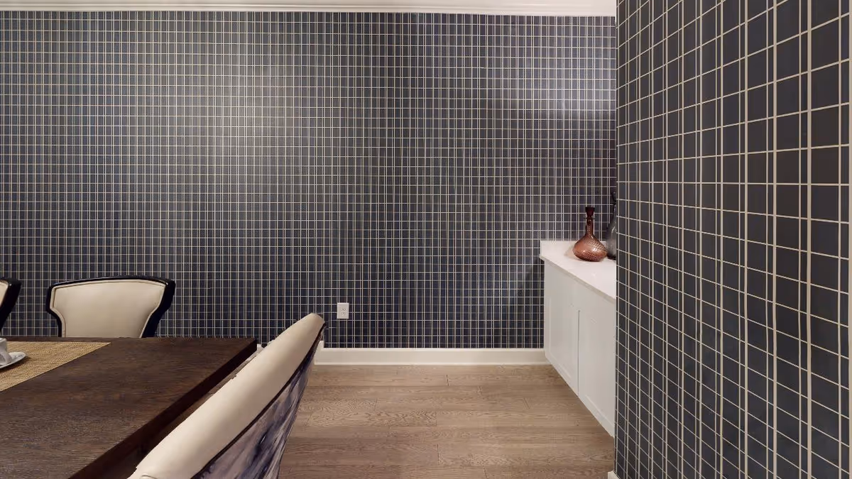 Interior view of a dining room with a dark wooden table and cream-colored chairs. The walls are covered with dark tiles featuring a light grid pattern. A white cabinet with decorative vases is visible against the wall.