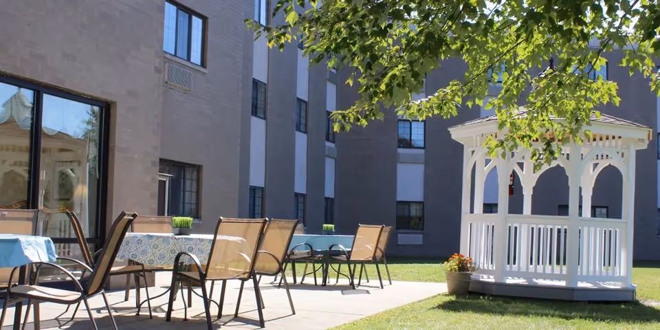 Outdoor patio area at Southwestern Nursing and Rehabilitation Center featuring several tables with chairs, a white gazebo, and a building with multiple windows in the background. Green grass and tree branches with leaves are visible.