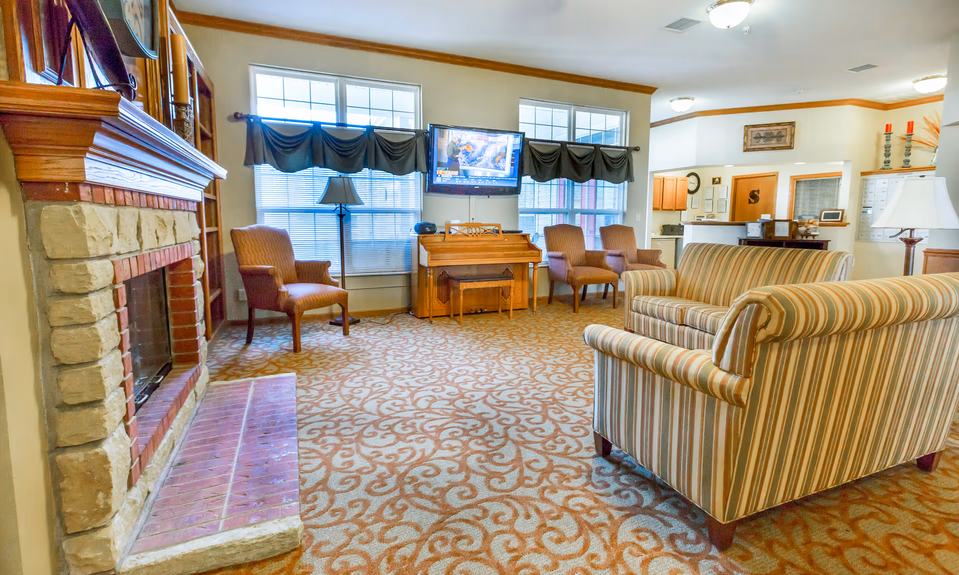 Cozy common room with striped sofas and chairs, a stone fireplace, wall-mounted TV, and large windows.