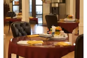 A dining area in Grandview Nursing Home featuring round tables covered with burgundy tablecloths, each set with yellow napkins, condiments, and small flower arrangements. The room has wooden flooring and comfortable upholstered chairs, with large windows allowing natural light to fill the space.