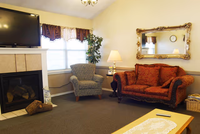 A cozy living room with a red patterned sofa, a gray armchair, a wooden coffee table with a lace runner, a fireplace with a TV mounted above it, a window with curtains, a floor lamp, a potted plant, and a large ornate mirror on the wall.