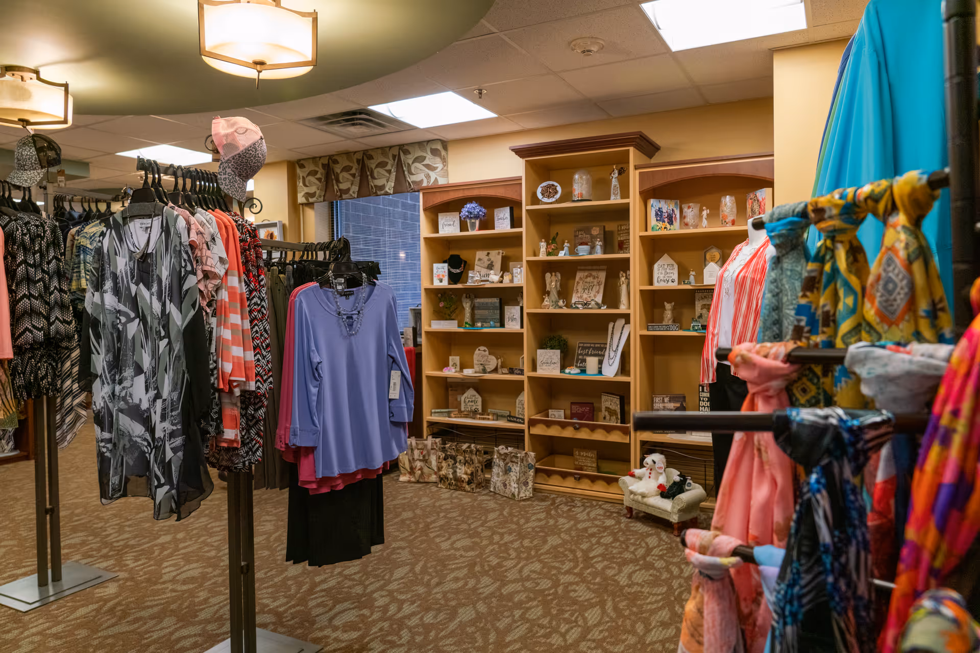 Interior view of a boutique or gift shop area within a senior living facility, featuring racks of women's clothing and scarves on display. In the background, wooden shelves hold various decorative items and small gifts. The space is well-lit with ceiling lights and has patterned carpet flooring.