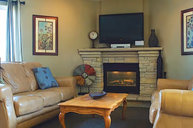 A cozy living room with two beige leather sofas, a wooden coffee table, a stone fireplace with a TV mounted above it, and framed floral artwork on the walls.