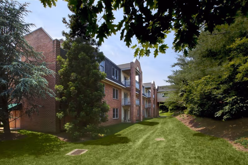 Brick three-story senior living building with balconies overlooking a grassy courtyard bordered by trees.