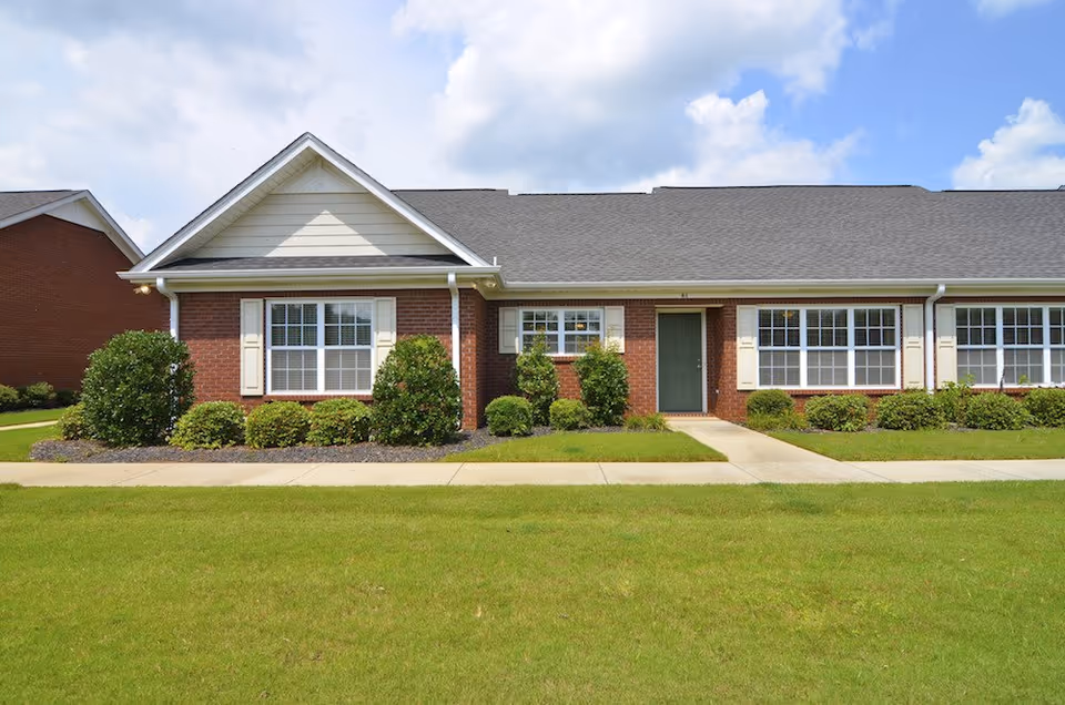 Exterior view of a single-story brick building with white trim, multiple windows with white shutters, a green door, and a well-maintained lawn and shrubs in front under a partly cloudy sky.