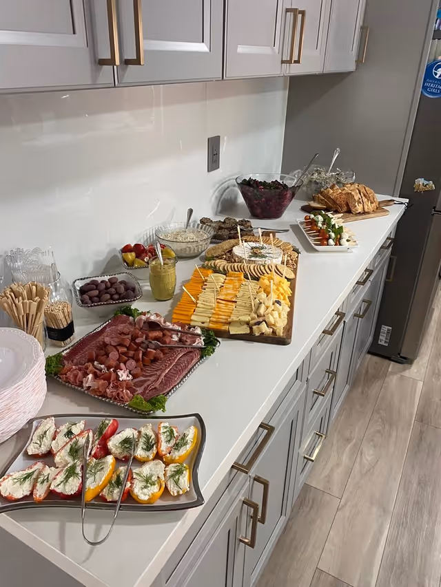 A kitchen counter with a variety of appetizers and snacks arranged on it, including sliced meats, cheese cubes, stuffed mini peppers, olives, bread slices, and a bowl of salad. The counter has white cabinets with gold handles and a light wood floor.
