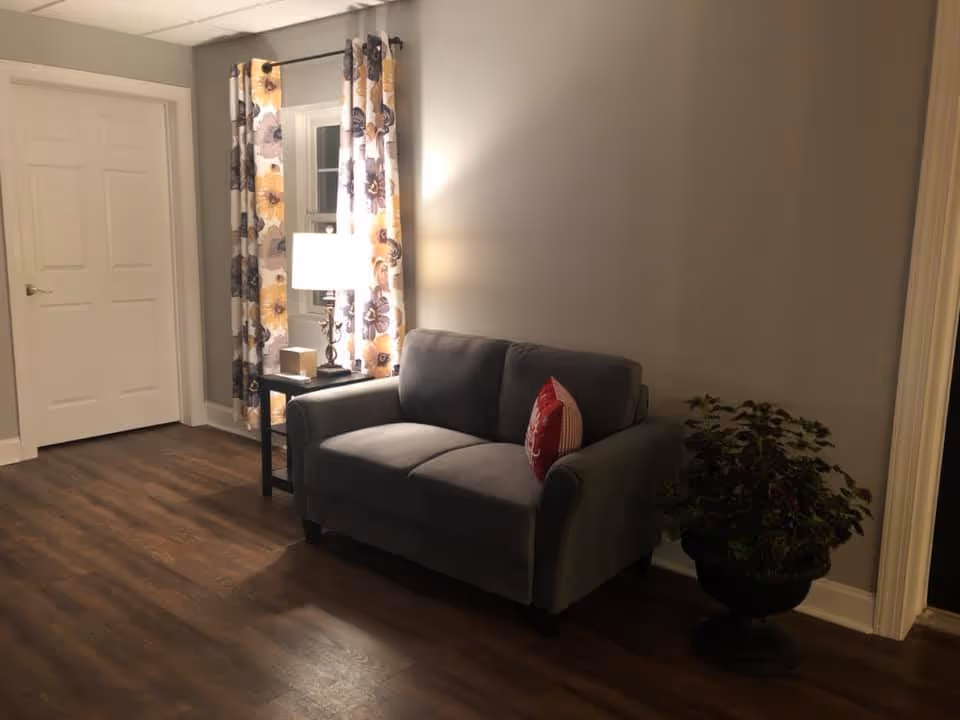 Small sitting area with a gray loveseat, side table and lamp by a curtained window and a potted plant.