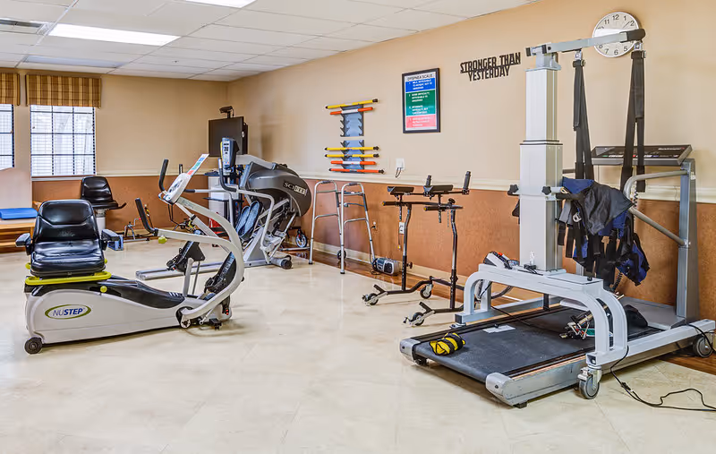 A rehabilitation or physical therapy room with exercise equipment including a recumbent bike, treadmill with harness, parallel bars, and other therapy tools. The room has beige walls, a window with blinds, and a motivational sign on the wall that reads 'STRONGER THAN YESTERDAY'.