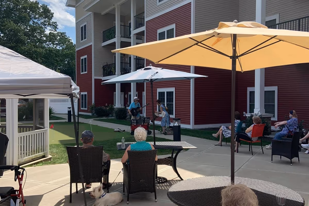 Outdoor patio area at Colebrook Village at Hebron with several elderly people seated on chairs under large umbrellas, watching a live musical performance by a man playing guitar and a woman singing. The scene includes a putting green, a small white dog, and a multi-story residential building with red and beige siding in the background.