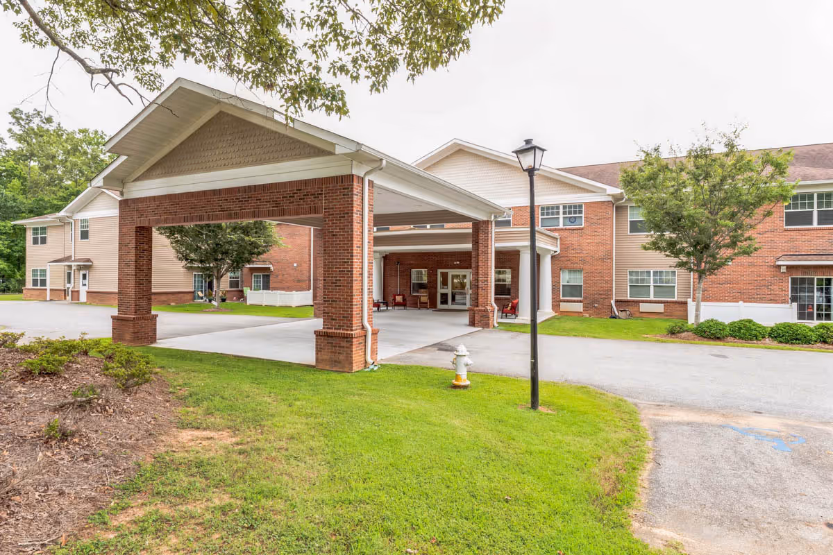Exterior view of a senior living facility building with a covered entrance supported by brick columns. The building has a combination of brick and beige siding, multiple windows, and a well-maintained lawn with trees and shrubs. A streetlamp and a fire hydrant are visible near the driveway.