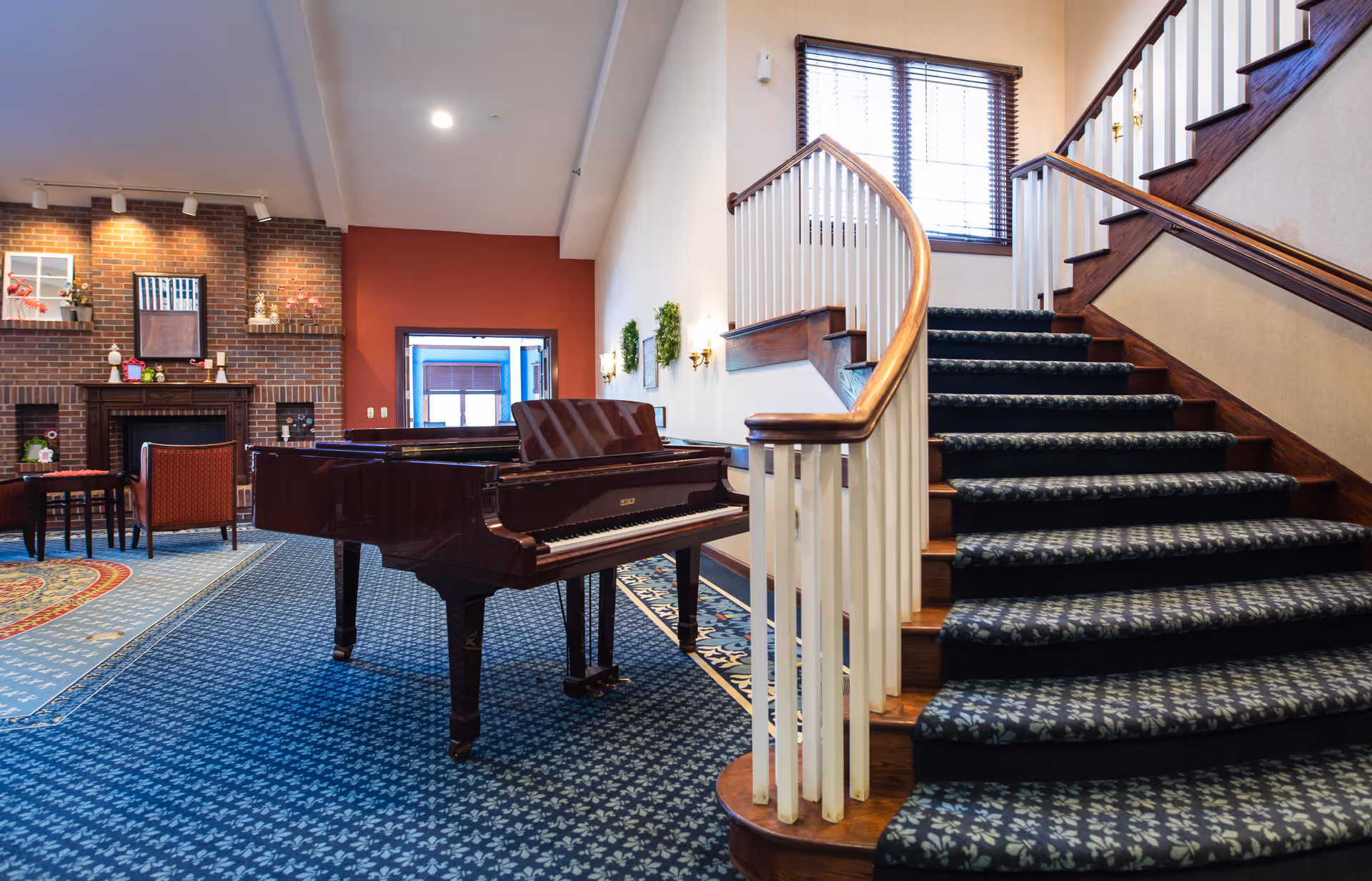 Interior view of a senior living facility common area featuring a grand piano on a patterned blue carpet, a curved staircase with wooden handrails and carpeted steps, a brick fireplace with decorative items on the mantel, and seating with red upholstered chairs. The walls are decorated with small green wreaths and there is a window with blinds near the staircase.