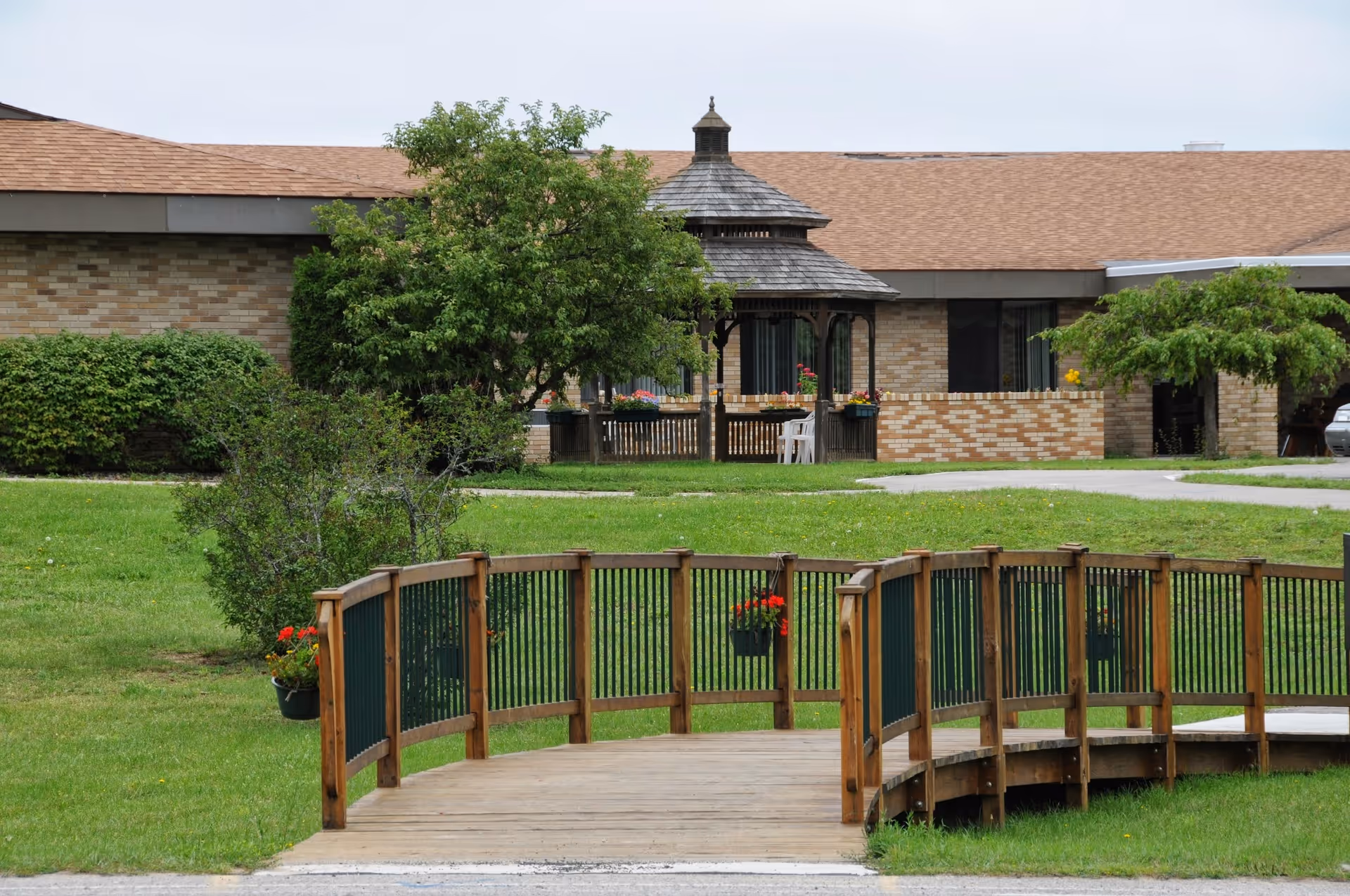 Outdoor view of Wellspring Lutheran Services Fairview Campus showing a wooden curved bridge over a grassy area, with a gazebo and brick building in the background surrounded by trees and shrubs.