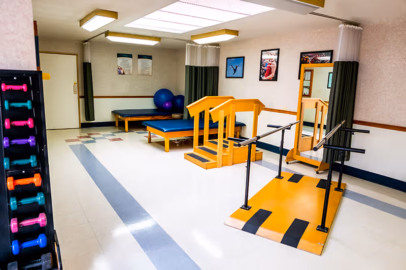 A physical therapy room with exercise equipment including colorful dumbbells on a rack, two blue padded therapy tables, large exercise balls, a wooden step platform with handrails, and a yellow ramp with parallel bars. The room has white walls with framed pictures and a large mirror, and fluorescent ceiling lights.