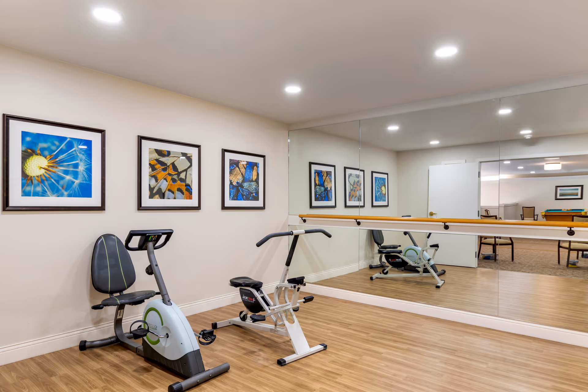 A small exercise room with two stationary exercise bikes on a wooden floor. The wall behind the bikes has three framed pictures of close-up nature scenes. A large mirror covers the adjacent wall, reflecting the room and part of another room with chairs and a pool table.