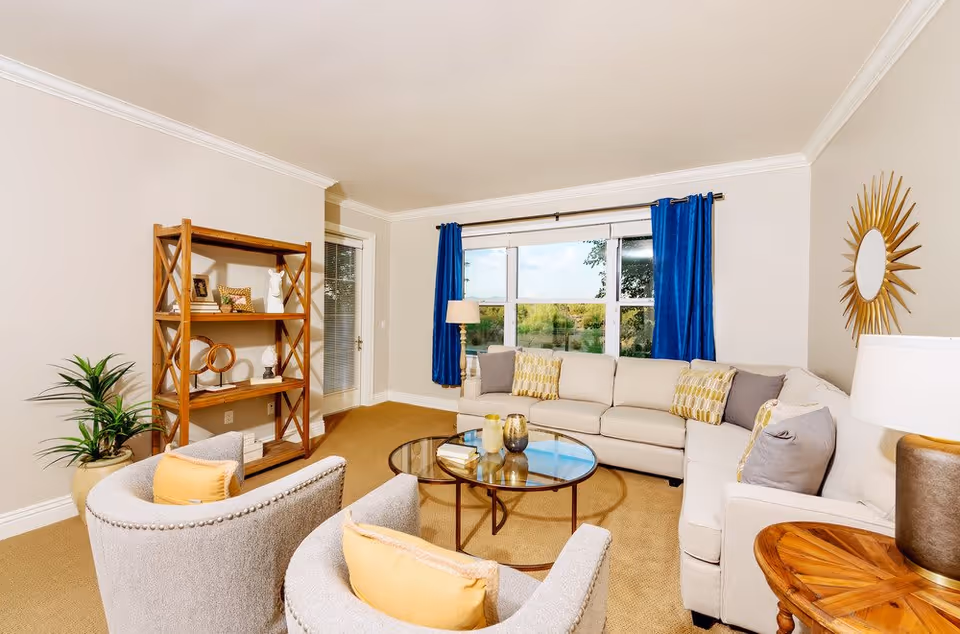 A bright and cozy living room with a large beige sectional sofa adorned with patterned and solid pillows. Two gray armchairs with yellow cushions face a round glass coffee table with decorative items. A wooden bookshelf with various decor pieces stands against one wall, next to a potted plant. The room features a large window with blue curtains, a floor lamp, a round wooden side table with a lamp, and a sunburst mirror on the wall.