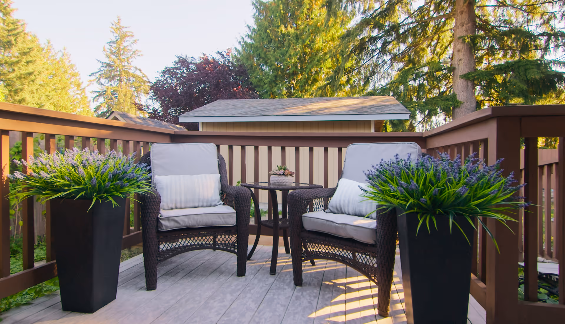 Outdoor patio area with two cushioned wicker chairs and a small round table between them, flanked by two tall black planters filled with green plants and purple flowers. The patio is surrounded by a wooden railing and trees are visible in the background.