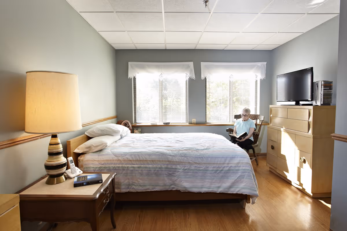 Sunny bedroom with a made bed, nightstand and lamp, dresser with TV, and a person seated reading by the windows.