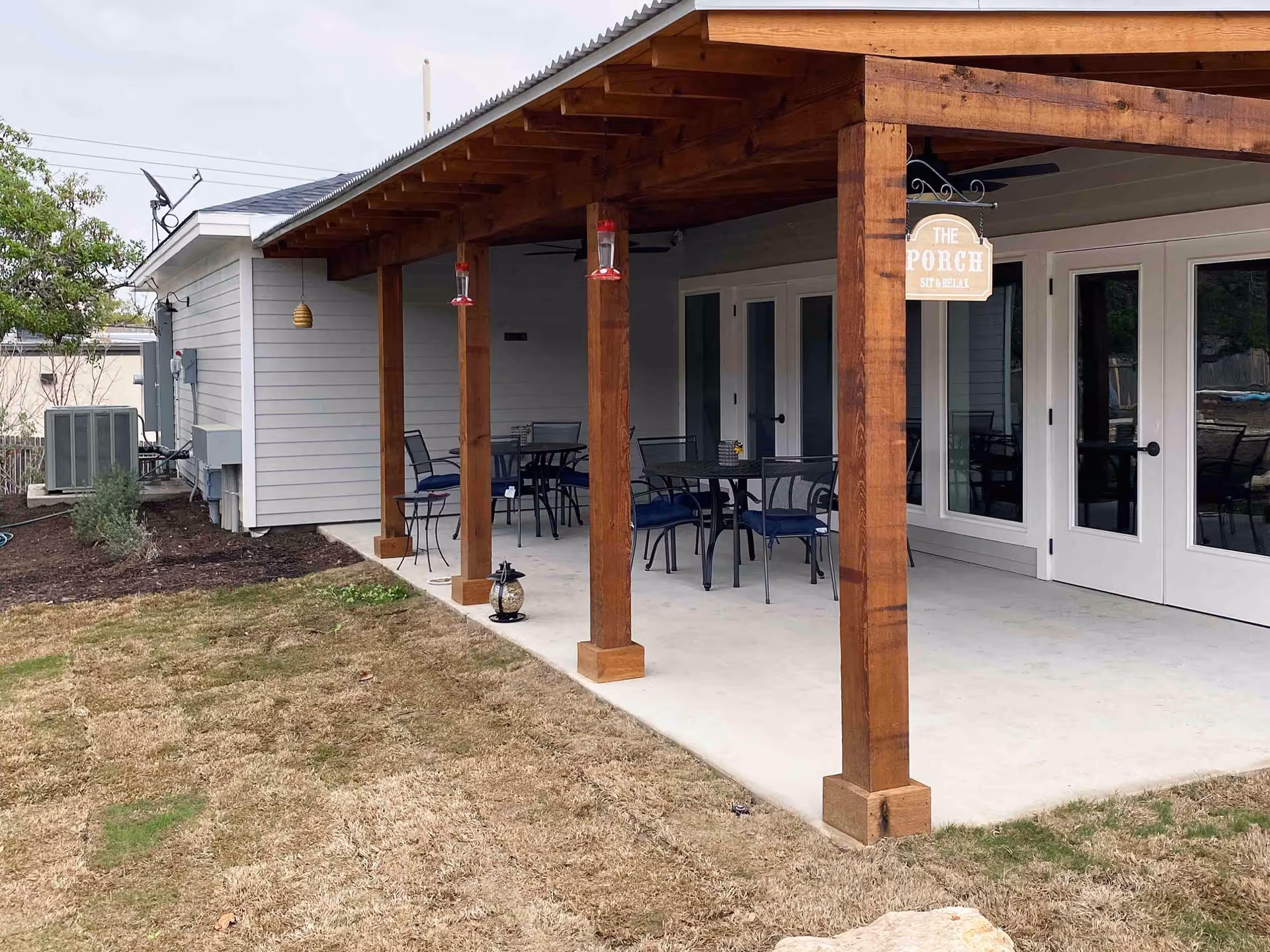 Outdoor covered patio area with wooden beams and ceiling fans at BeeHive Homes of Crownridge Assisted Living. The patio has several tables and chairs arranged for seating. A sign hanging from one of the beams reads 'The Porch Sit & Relax'. The patio opens to a grassy yard with some landscaping and an air conditioning unit visible on the side of the building.