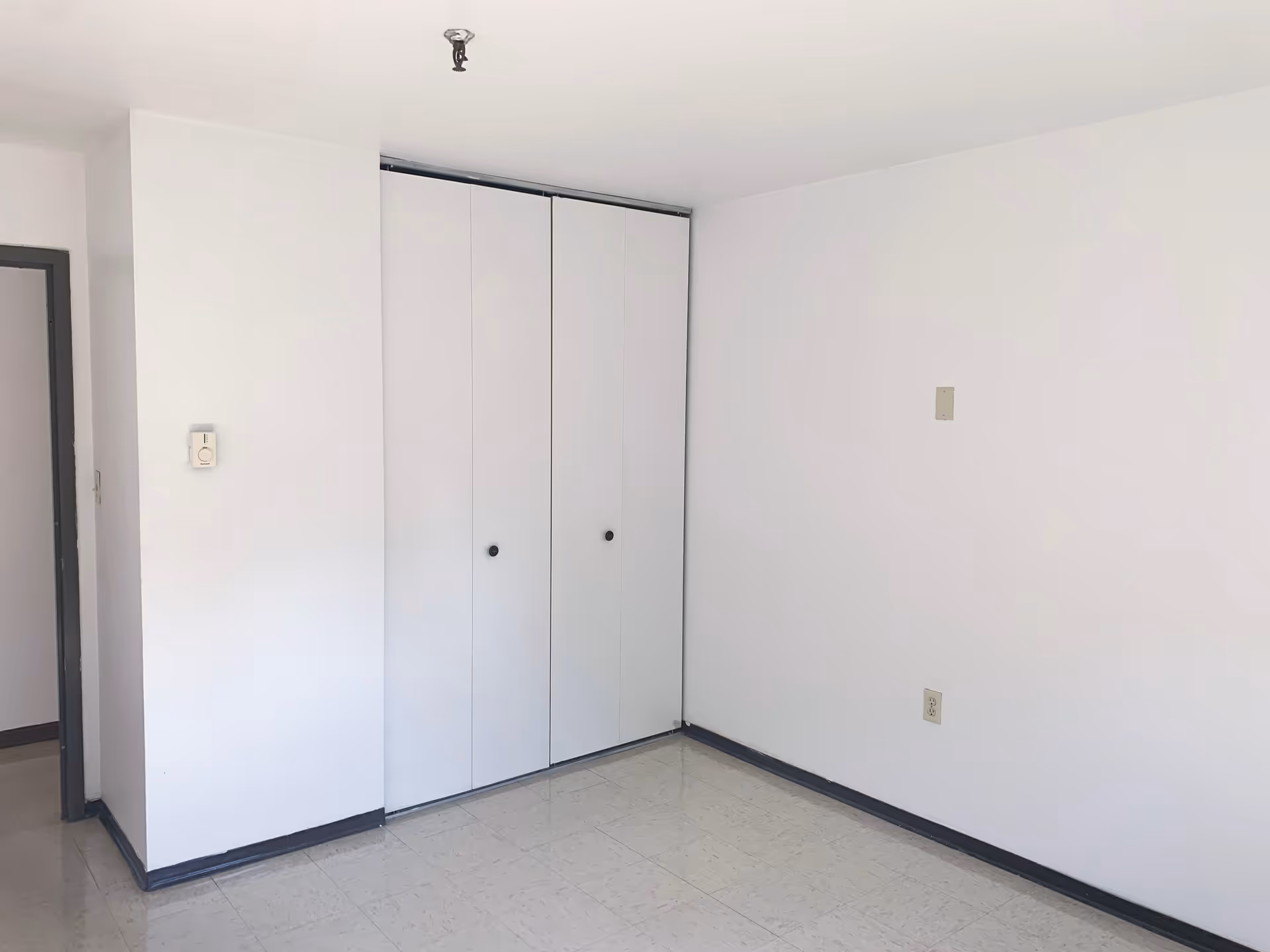 Empty white room with closed closet doors, linoleum tile floor, and a bare ceiling light fixture.