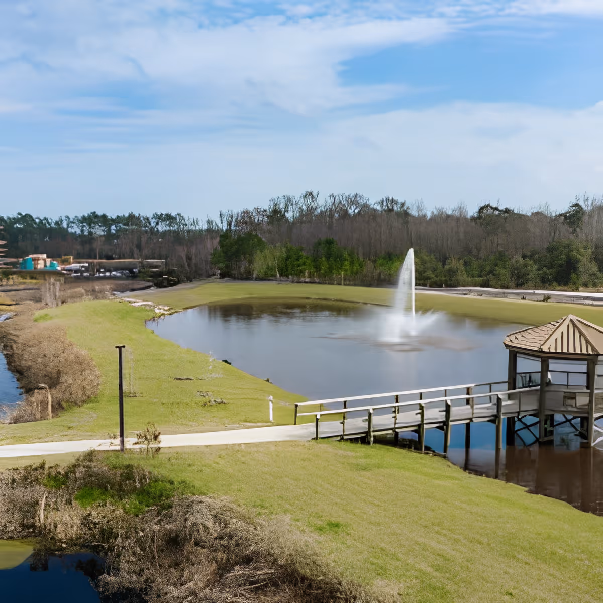 View of a landscaped pond with a central fountain, surrounding grassy lawns and a wooden gazebo on a boardwalk.