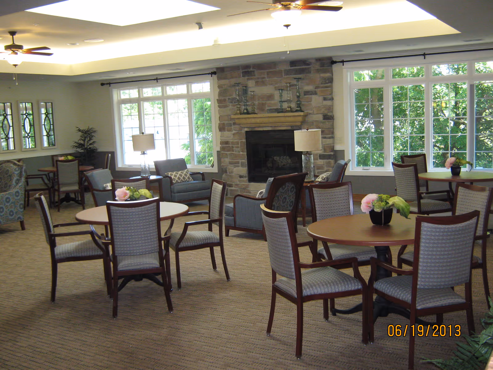 Bright communal room with round tables and chairs, a seating area, stone fireplace and large windows.