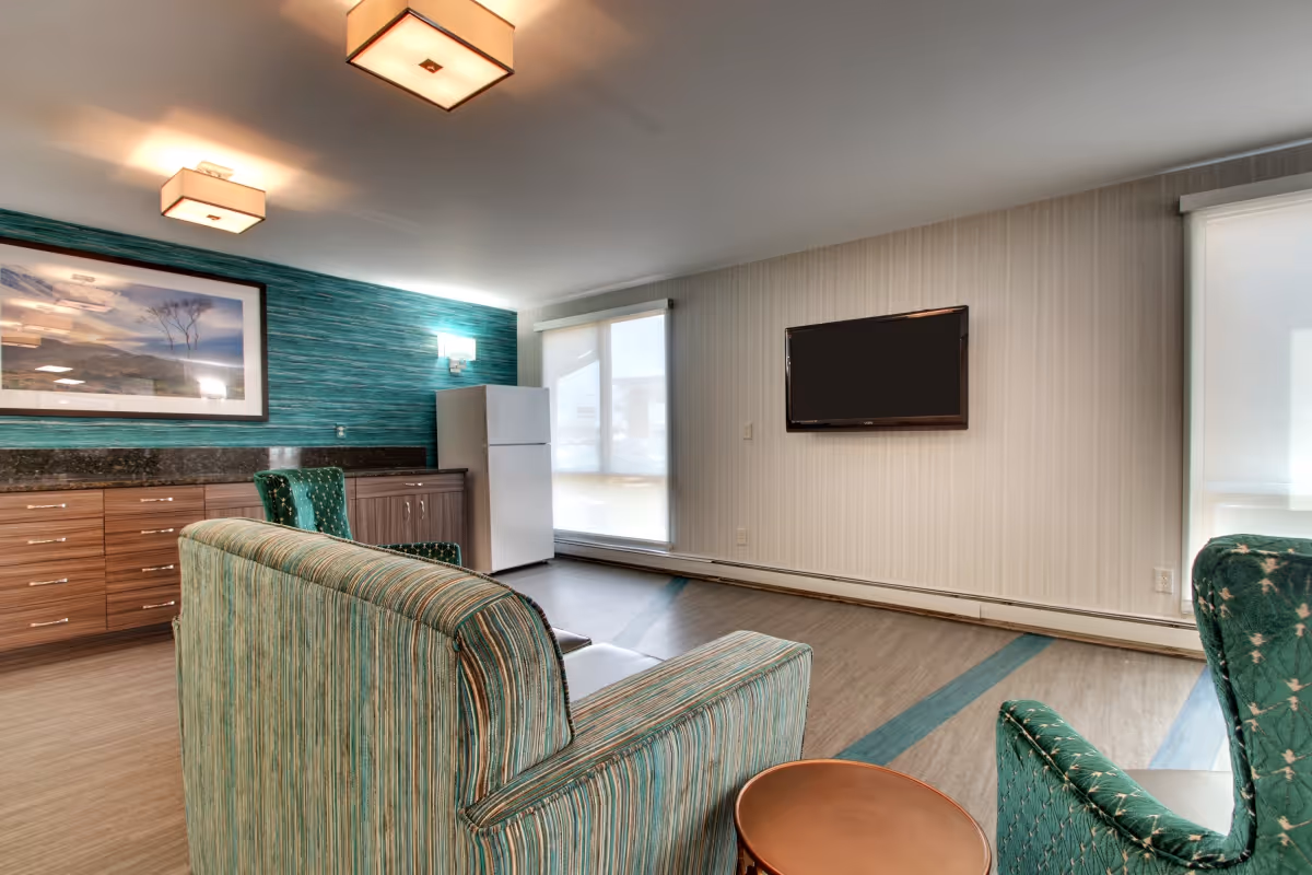A common area in a senior living facility featuring a striped green sofa and two green armchairs around a small round table. The room has a mounted flat-screen TV on a light-colored wall, a white refrigerator, wooden cabinets with a dark countertop, and a large framed landscape photograph on a teal accent wall. Two large windows with white blinds allow natural light into the space.