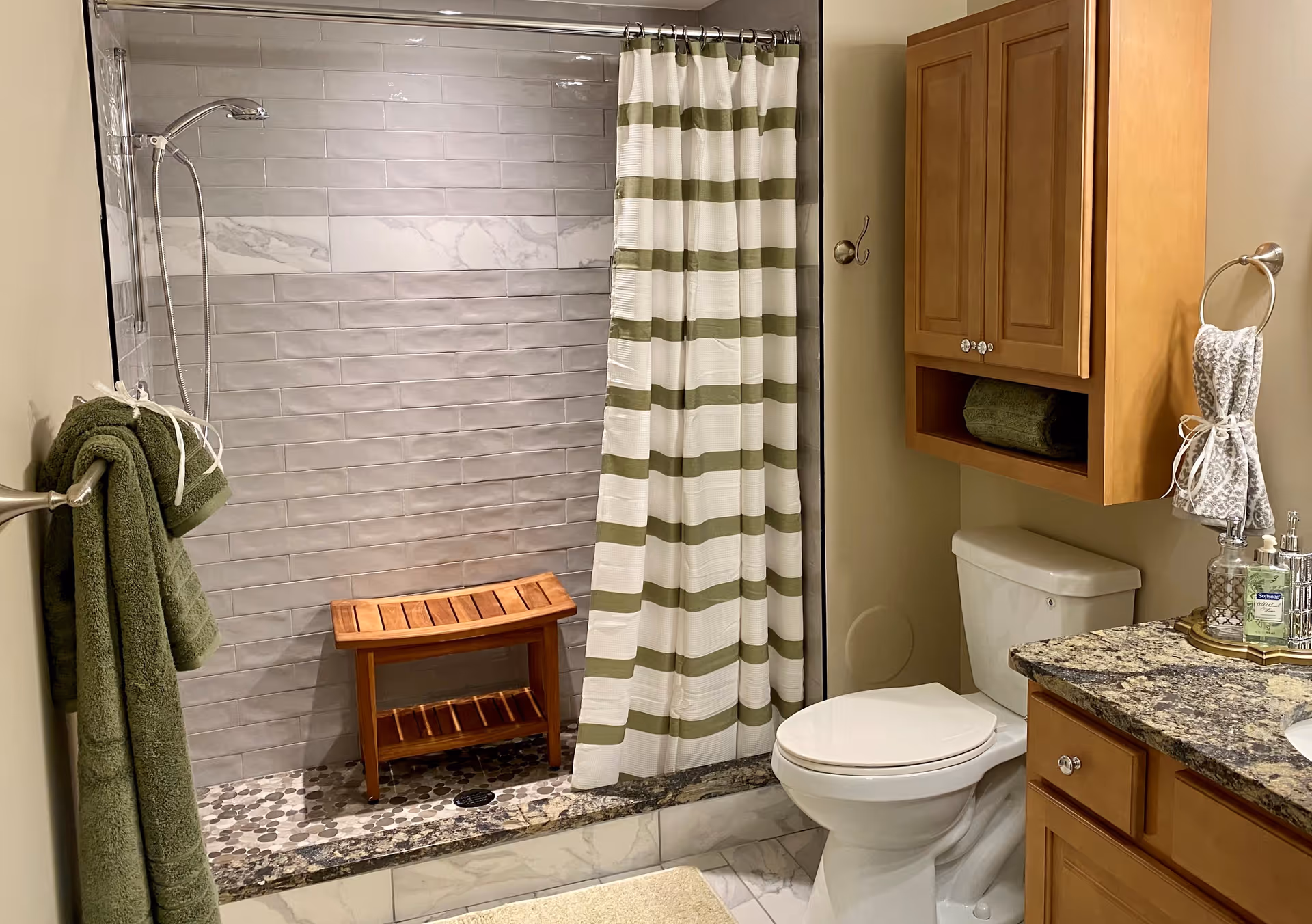 A bathroom with a walk-in shower featuring gray subway tiles and a decorative marble tile strip. The shower has a wooden bench and a green and white striped shower curtain. There is a white toilet next to a wooden cabinet mounted on the wall. A granite countertop with a sink is partially visible, along with a towel ring holding a patterned hand towel and a soap dispenser. Green towels hang on a towel bar on the left wall.