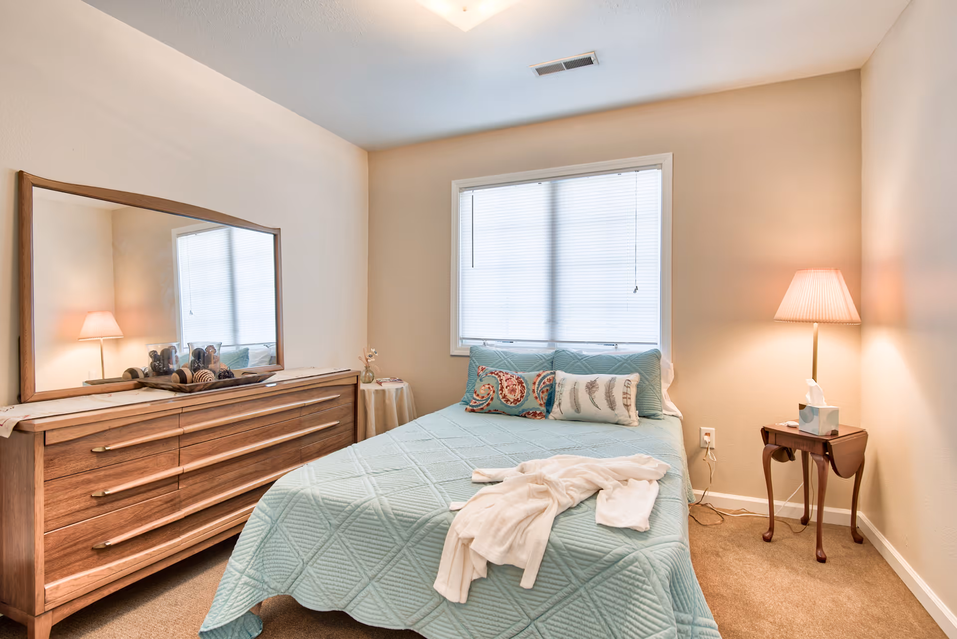 A cozy bedroom with a bed covered in a light blue quilt and decorative pillows. A white robe is laid out on the bed. To the left is a wooden dresser with a large mirror and decorative items on top. A small round table with a vase and a lamp on a side table are also visible. The room has beige walls and carpeted floor with a window covered by blinds.