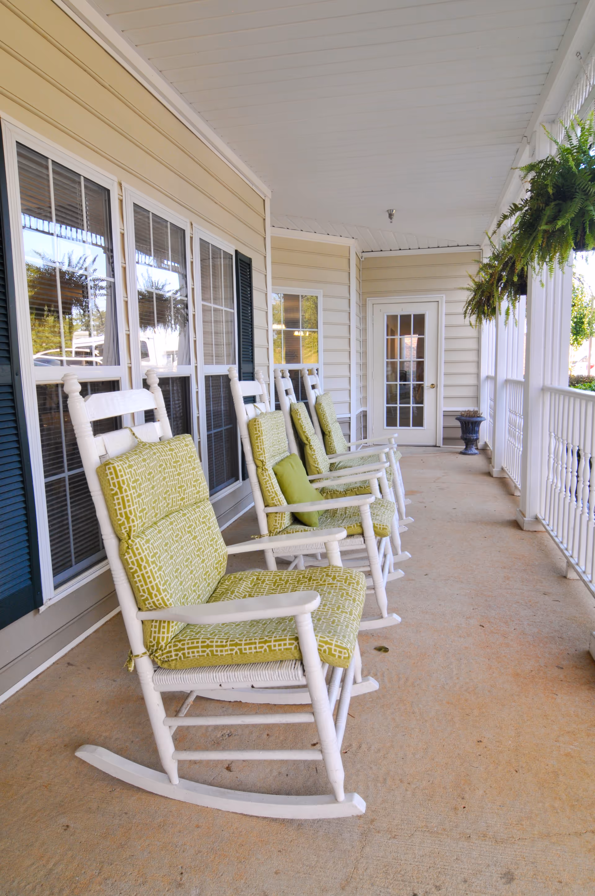 A covered porch area with a row of white wooden rocking chairs featuring green patterned cushions and small green pillows. The porch has beige siding, multiple windows with dark shutters, a white ceiling, and hanging green plants. A white door with glass panes is visible at the end of the porch.
