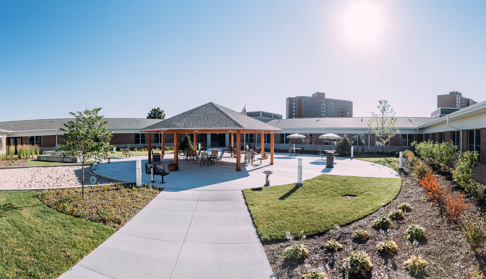 Outdoor courtyard area of a senior living facility with a central covered pavilion featuring tables and chairs. Surrounding the pavilion are landscaped gardens, a sand volleyball court, and a paved walkway. The building forms a U-shape around the courtyard under a clear blue sky with the sun shining brightly.