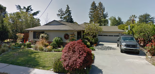 Single-story house with a gray roof and beige exterior surrounded by trees and shrubs. A black SUV is parked on the concrete driveway leading to a two-car garage. The front yard has a well-maintained lawn, various plants, and a small tree with red leaves near the driveway.