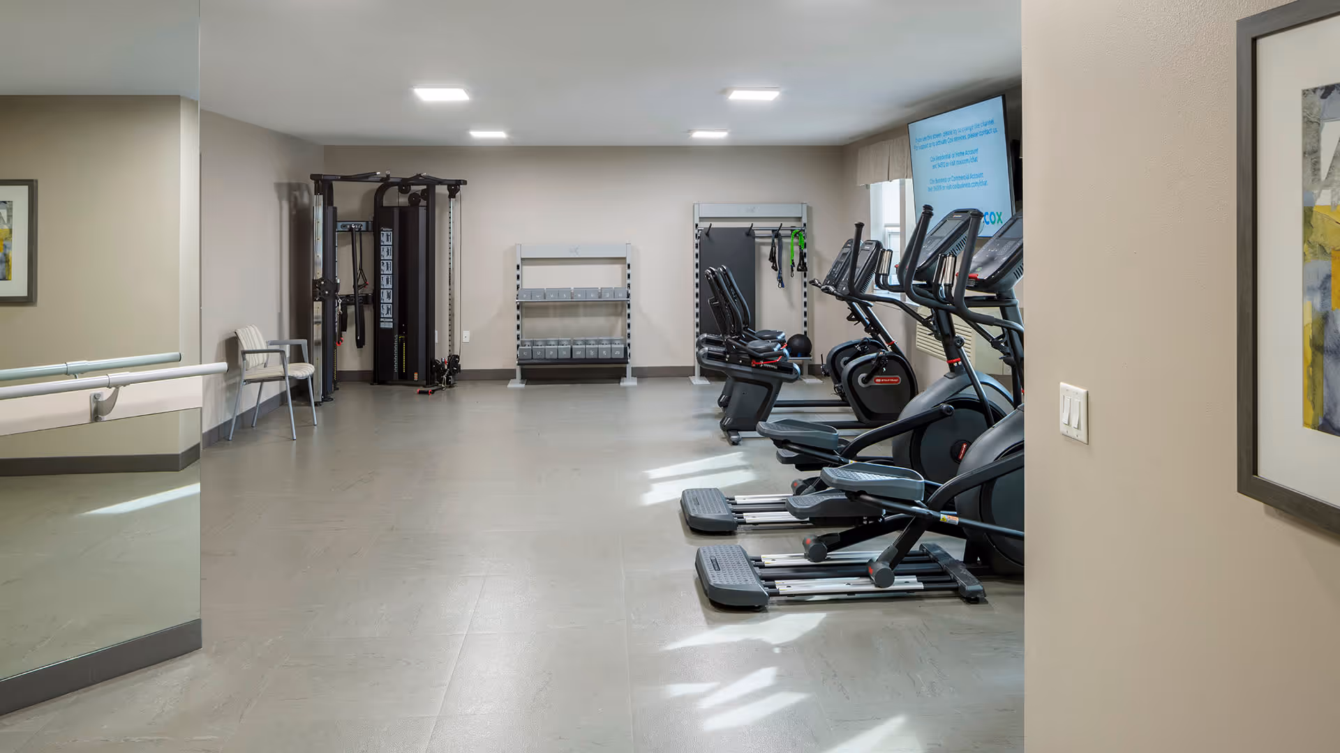 A fitness room with exercise equipment including elliptical machines, recumbent bikes, a weight rack with dumbbells, and a multi-functional cable machine. The room has beige walls, a large mirror on the left wall, and a framed abstract painting on the right wall. There is a TV mounted on the far right wall above the exercise machines.
