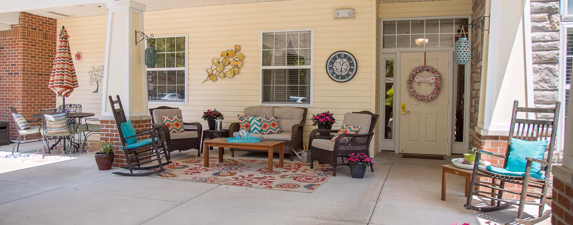 Covered outdoor patio area with wicker chairs, a loveseat, and rocking chairs arranged around a wooden coffee table on a decorative rug. The wall behind features two windows, a clock, and a door with a wreath. There are potted plants and colorful cushions on the seating. A small round table with chairs and a striped umbrella is visible to the left.