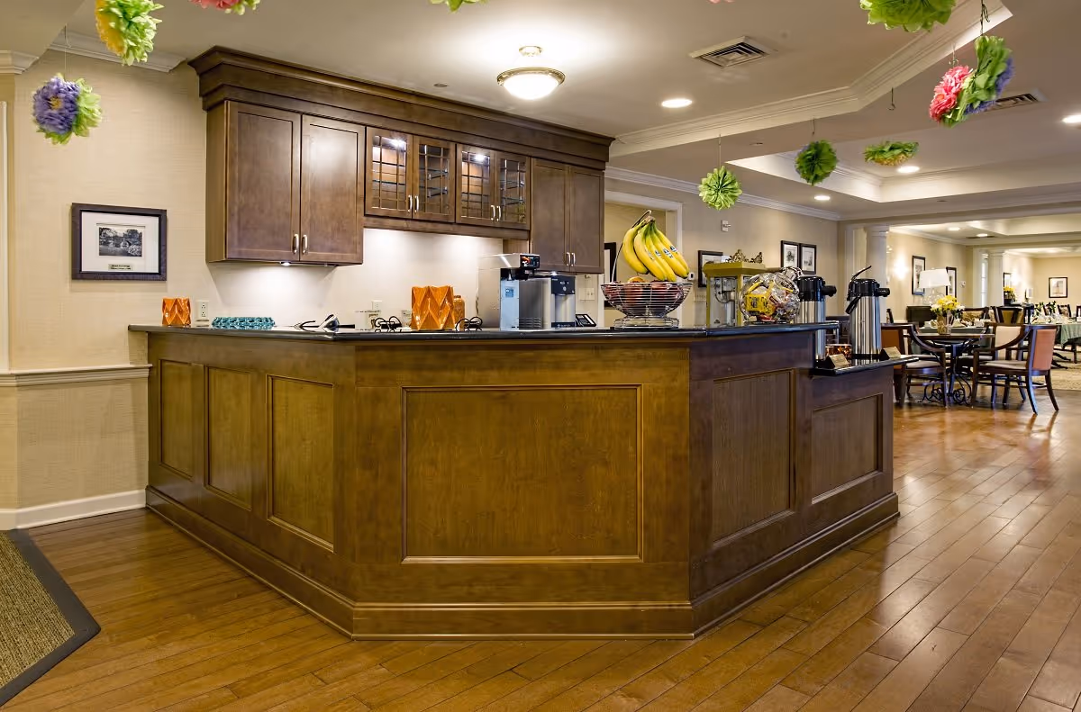 Interior view of a senior living facility's kitchen and dining area with a wooden counter, coffee machine, fruit basket with bananas, and tables with chairs in the background. Colorful hanging decorations are visible from the ceiling.