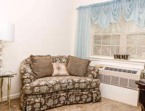 A floral-upholstered loveseat with decorative pillows sits next to a lamp under a window with a blue valance and an air-conditioning unit.