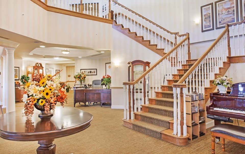 Interior view of a senior living facility lobby featuring a wooden staircase with white spindles and a carpet runner. A round wooden table with a large floral arrangement is in the foreground. To the right, there is a piano with a floral arrangement on top and a piano bench. In the background, a wooden desk with a chair and more floral arrangements is visible. The walls are decorated with framed pictures and the space is warmly lit.