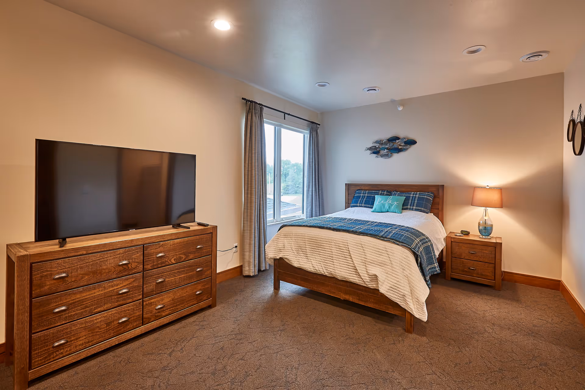 A cozy bedroom with a wooden bed frame, white and blue bedding, and a decorative pillow that says 'Relax'. There is a wooden dresser with a large flat-screen TV on top, a window with curtains, a wooden nightstand with a lamp, and wall decorations including a school of fish sculpture and two round mirrors.