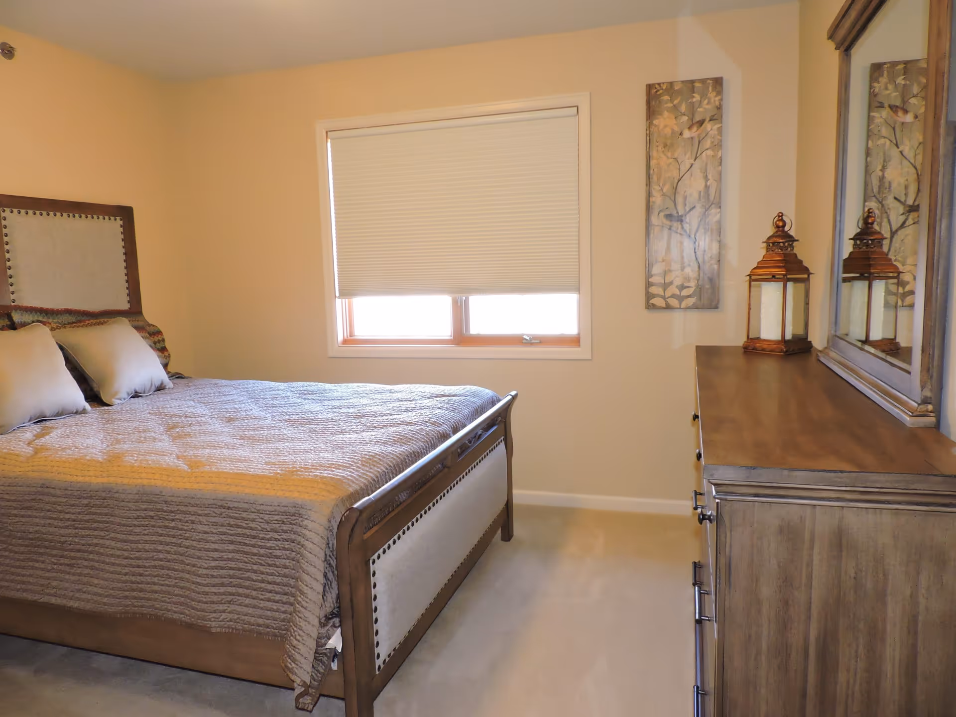 Furnished bedroom with a made bed, window with a closed shade, dresser and mirror and wall decor.