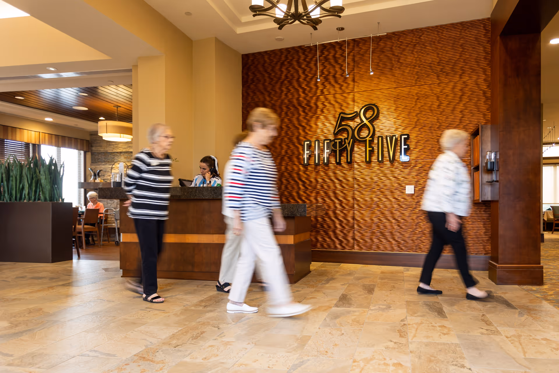 Interior view of a senior living facility lobby area with a reception desk and a textured wall displaying the number 58 and the words 'Fifty Five'. Several elderly women are walking through the space, and a staff member is seated behind the reception desk. The area has tiled flooring, warm lighting, and modern decor.
