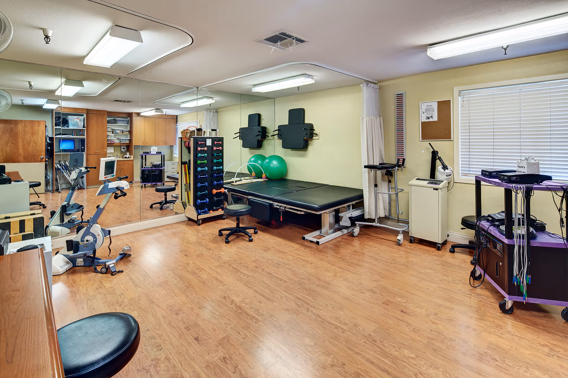 A rehabilitation or physical therapy room with wooden flooring, a large wall mirror, exercise equipment including stationary bikes, a rack of colorful dumbbells, two green exercise balls, a black therapy table, and various medical devices. The room has a window with blinds and light yellow walls.