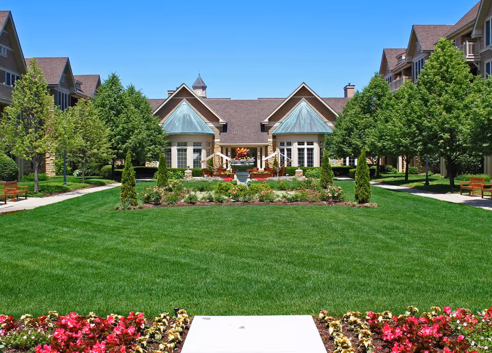 A well-maintained outdoor garden area with lush green grass, colorful flower beds, and neatly trimmed trees. The garden is flanked by benches and pathways on both sides, leading to a building with peaked roofs and large windows under a clear blue sky.