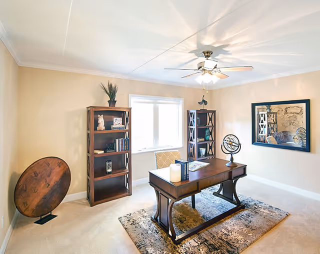 A bright and spacious home office with beige walls and carpet. The room features a wooden desk with decorative items including a candle, books, and a globe. Behind the desk are two wooden bookshelves filled with books and decorative pieces. A large window lets in natural light, and a ceiling fan with lights is mounted on the ceiling. A large framed mirror hangs on one wall, and a round wooden decorative piece stands on the floor in the corner.