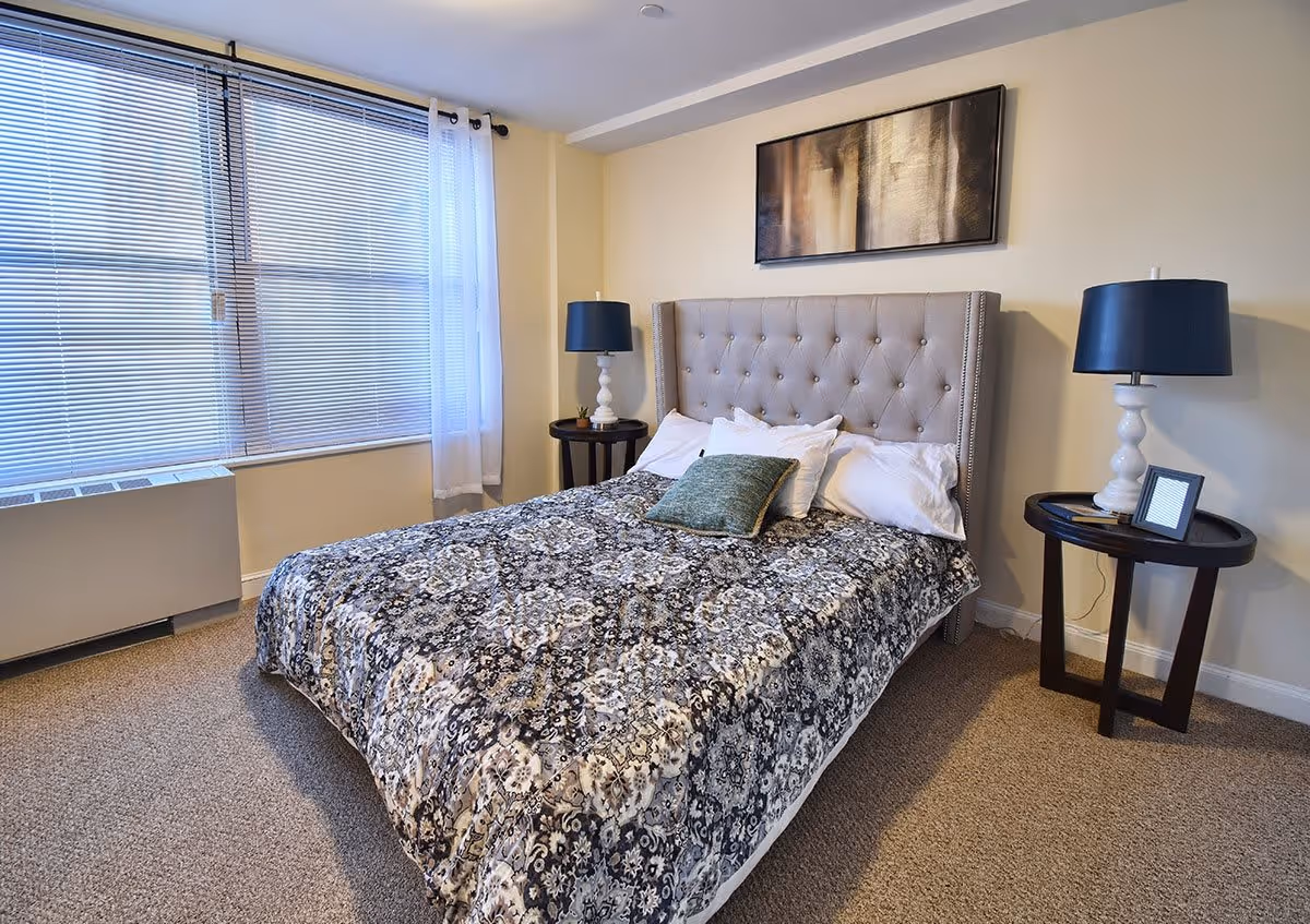 A neatly made bed with a patterned comforter and several pillows in a bedroom. The bed has a tufted headboard and is flanked by two round bedside tables, each with a lamp. A framed abstract painting hangs above the bed. A large window with blinds and sheer curtains is on the left side of the room.