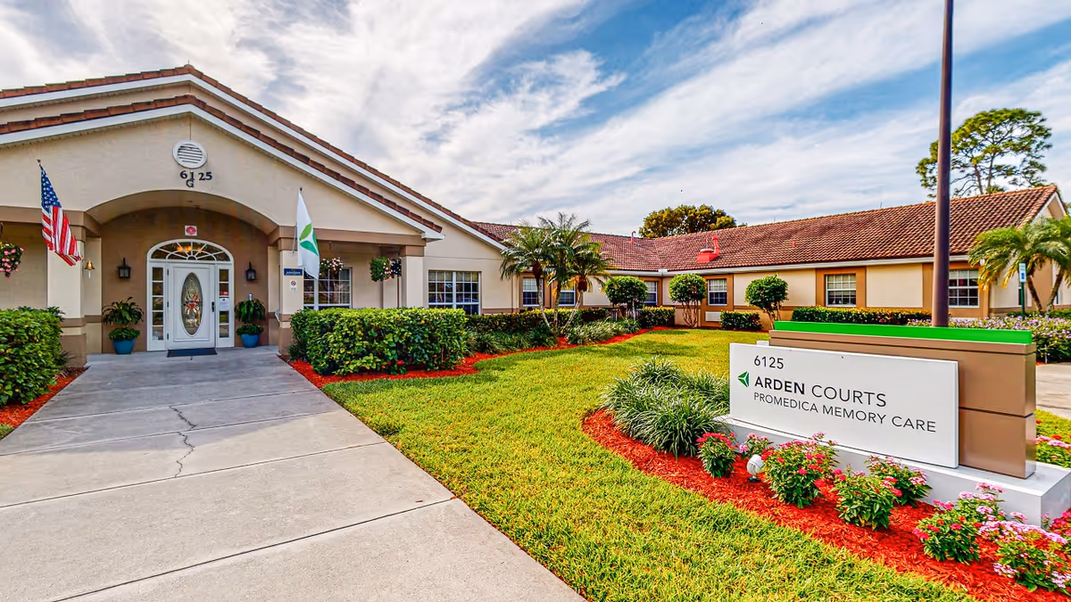 Exterior view of Arden Courts ProMedica Memory Care building with a walkway leading to the entrance, surrounded by well-maintained green lawns, bushes, and flowers. The building has a beige facade with a tiled roof, and there are two flags near the entrance.