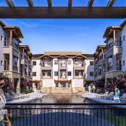 View of a senior living facility courtyard with a central water feature and fountain, surrounded by multi-story buildings with balconies. There are lounge chairs and flowering bushes along the sides, and several people walking or standing around the area under a pergola structure.