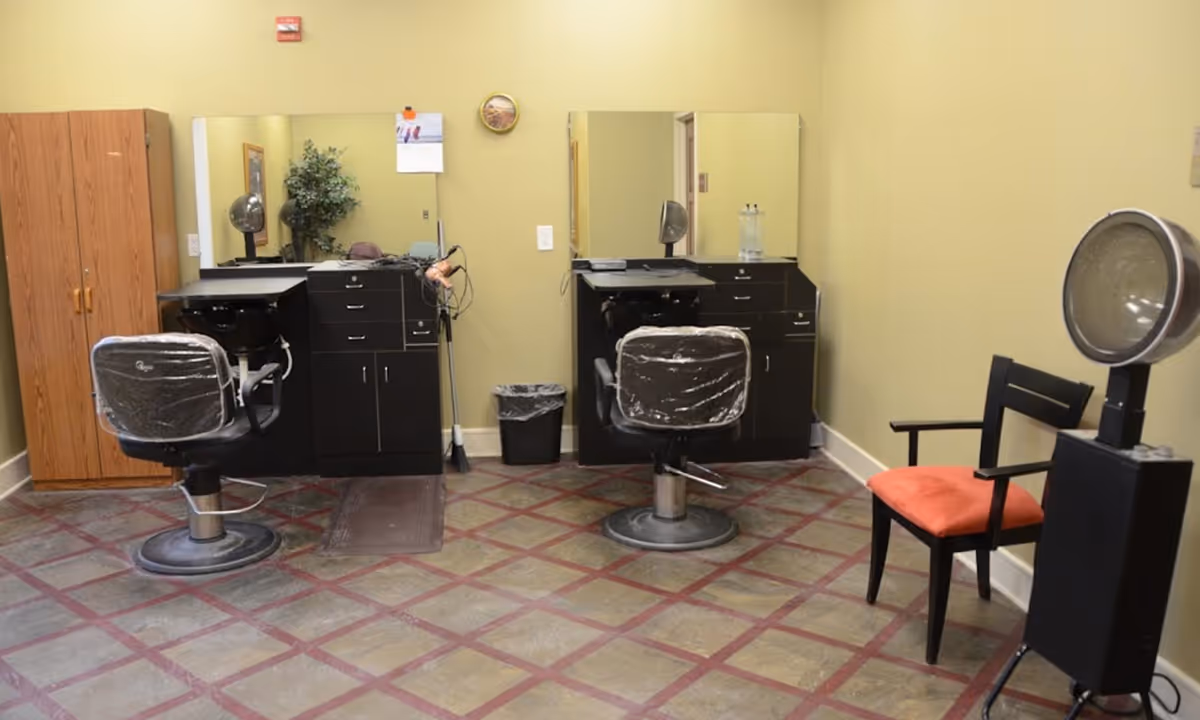 Small salon room with two styling stations, chairs, mirrors, cabinets and a hooded hair dryer.