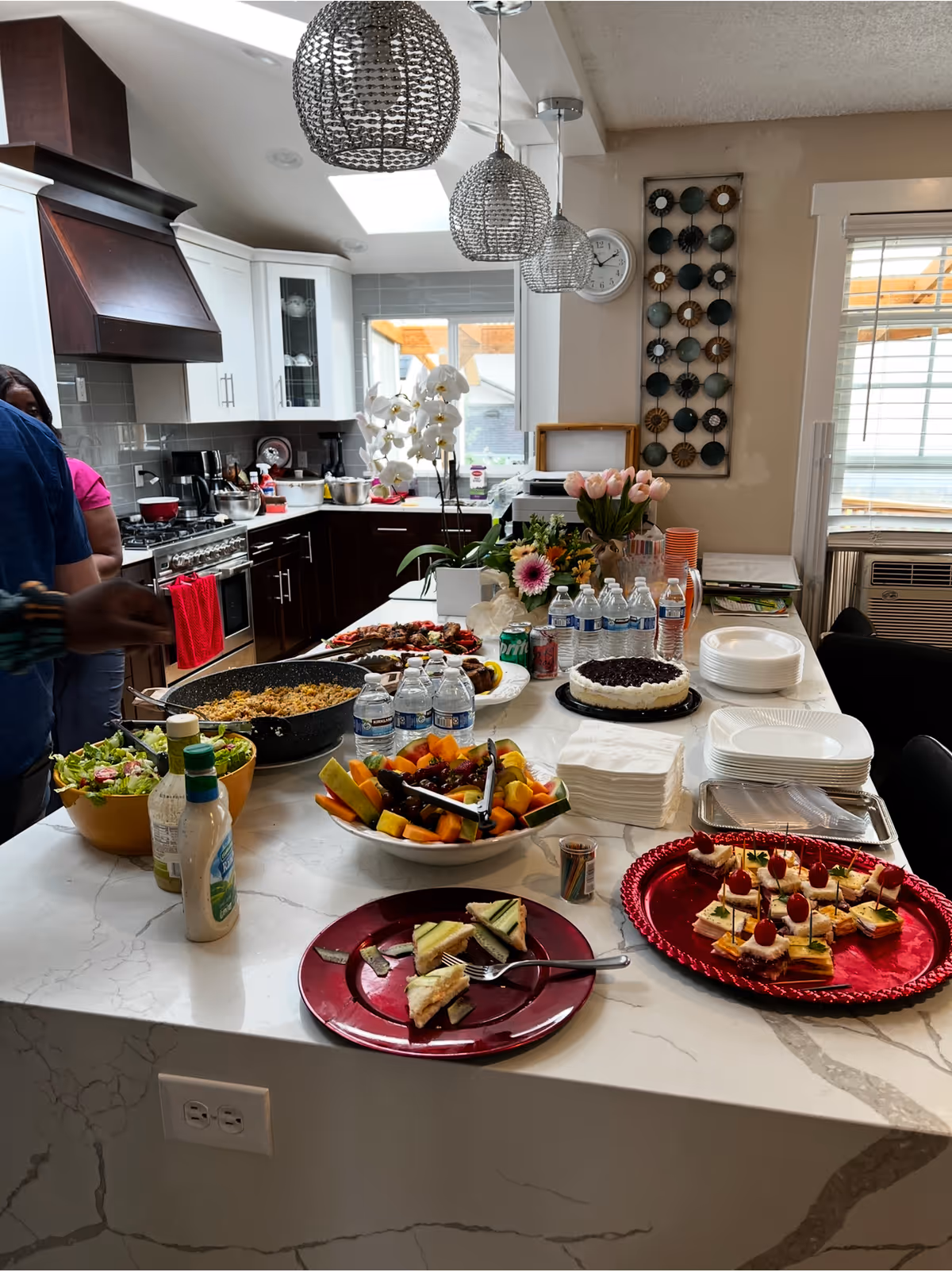 A modern kitchen with a large white marble island countertop set with various foods including a bowl of salad, a bowl of mixed fruit, a pan of cooked rice, bottled water, a cake, and plates of finger sandwiches and appetizers. There are three decorative pendant lights hanging above the island. In the background, there are white and dark brown cabinets, a stove with a red towel, a window, and some decorative flowers and wall art. Two people are partially visible on the left side of the image.