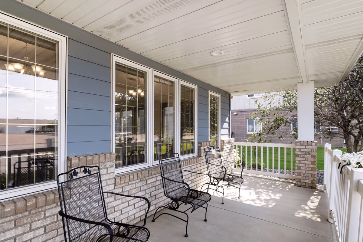 Covered outdoor patio area with three black metal chairs lined up against a brick wall with large windows. The patio has a white railing and a white ceiling, with some greenery and a tree visible outside.