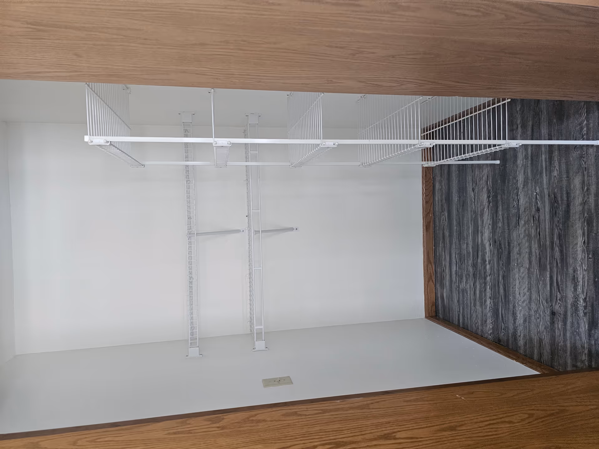 Empty closet with white wire shelving on the back wall and a white wire rack on the right side. The floor has dark wood-like vinyl flooring and the walls are painted white. The closet door frame is wooden.