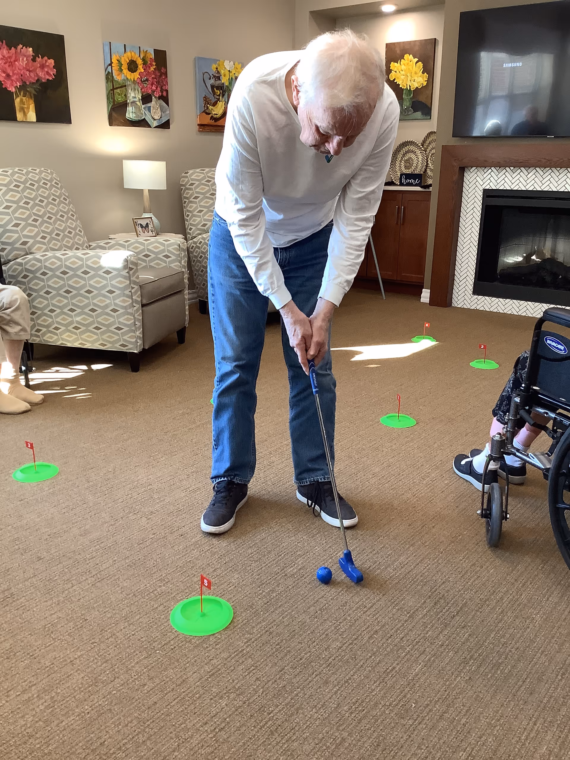 An elderly man playing indoor mini golf in a living room setting with patterned armchairs, floral paintings on the wall, a fireplace, and a television. There are several small green putting cups with red flags placed on the carpeted floor. Another person in a wheelchair is partially visible on the right side.